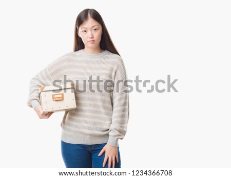 Young Chinese woman over isolated background holding a box with a confident expression on smart face thinking serious