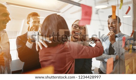 Two smiling young businesswomen hugging while brainstorming with sticky notes on a glass wall with colleagues in a modern office Royalty-Free Stock Photo #1234428997