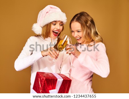 Christmas,x-mas, concept.Two smiling beautiful women in white winter sweaters.Girls posing on golden background.Models with big gift box drinking champagne in glasses.Having fun, celebrating New Year