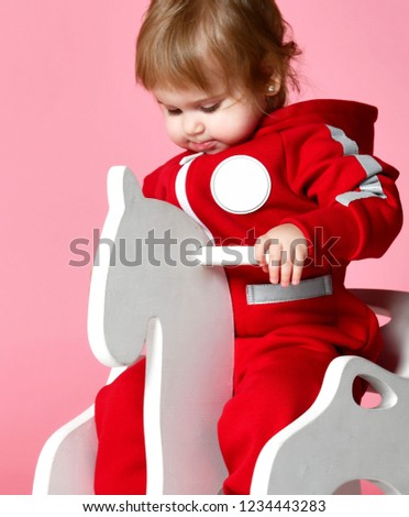 Toddler baby girl is riding swinging on a rocking chair toy horse over light pink background