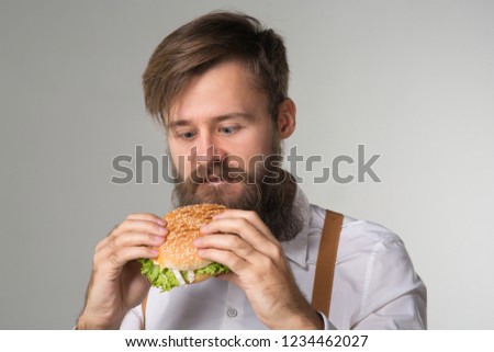 Man with beard in white shirt and suspenders eating junk food from a fast food hamburger or cheeseburger on gray background