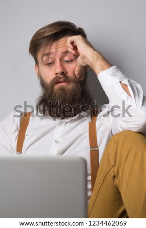 Man with beard in white shirt and suspenders works and communicates in a laptop on gray background