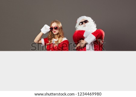 Cool Santa Claus and young beautiful mrs. Claus in sunglasses stand behind a white canvas on the gray background.
