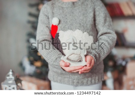 Girl standing at the Christmas tree and holding a toy horse. She's wearing a gray sweater. Christmas concept