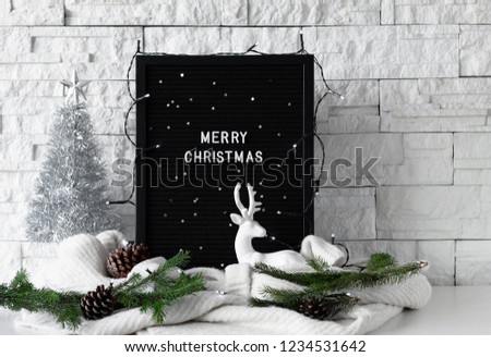 Merry Christmas text made with white letters on a black board with knitted sweaters and winter home decorations on white table and white background, side view