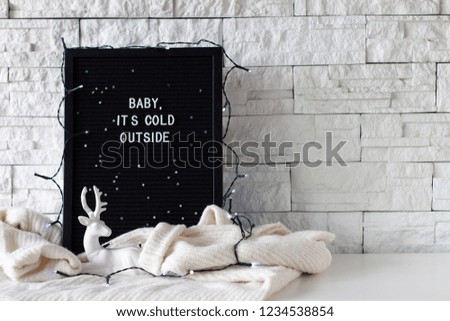 Baby it's cold outside phrase made with white letters on a black wooden desk with lights and with a deer statuette on white background