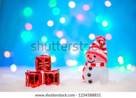 Christmas holiday picture with a cute little snowman in a red knitted hat and gifts in a red package in the snow on a blue background with lights
