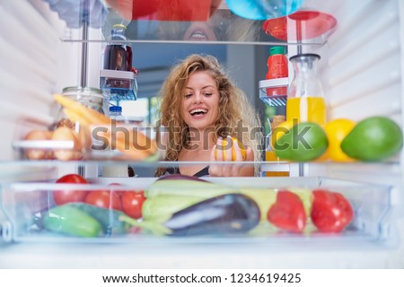 Woman standing in front of opened fridge and taking orange fruit. Fridge full of groceries. Picture taken from the iside of fridge.
