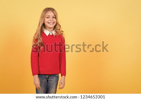 Child girl with long curly blond hair happy smile in red sweater and blue jeans on orange background. Fashion, style, beauty, look concept, copy space