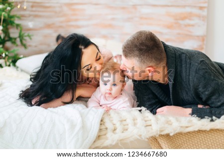 happy parents lying on the bed with their baby and kissing a girl in a head. Christmas tree by the bed.