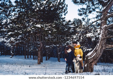 Happy european young family with big dog posing against winter pine forest. Horizontal family photo near a beautiful big old curve of pine.