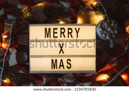 Merry Xmas text displayed on a lightbox with beautiful Christmas decoration with fir branches, red plaid and lights in the dark. Flat lay. Top view