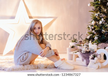 Beautiful young blond woman dressed in white sweater posing under Christmas tree in a holiday interior