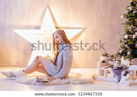 Beautiful young blond woman dressed in white sweater reading book under Christmas tree in a holiday interior