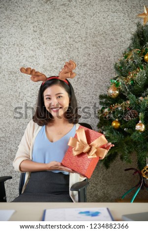 Portrait of joyful young Vietnamese business lady posing with Christmas present at office table