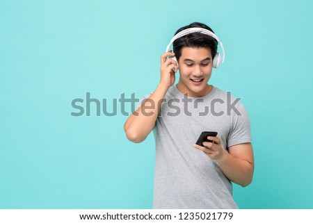 Happy young Asian man wearing wireless headphones listening to music from smartphone studio shot isolated on light blue background Royalty-Free Stock Photo #1235021779