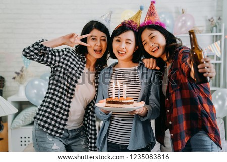 young cheerful woman blinking eyes face camera posing taking picture in decorated room for birthday house party. attractive ladies holding cake with candles on fire and beer wearing colorful hats.