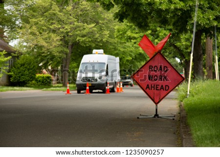 Orange and Black "Road Work Ahead" Sign in Residential Neighborhood with Work Vehicle and Orange Cones in Background, Daytime
