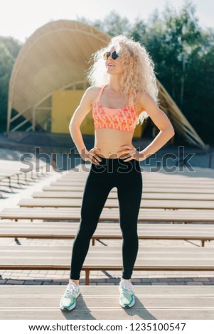 Photo of curly-haired athletic woman standing in park among shop