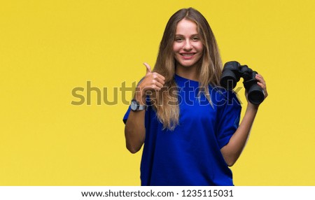 Young beautiful blonde woman looking through binoculars over isolated background happy with big smile doing ok sign, thumb up with fingers, excellent sign
