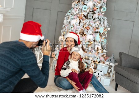 New Year. Christmas. Family. Mom and daughter are posing at camera while dad is taking pictures. All in Santa hats near the Xmas tree at home