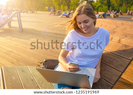 Happy smiling caucasian woman reading pleasant text message in messenger via mobile phone while sitting with portable laptop computer in park in sunny summer day during leisure time. 