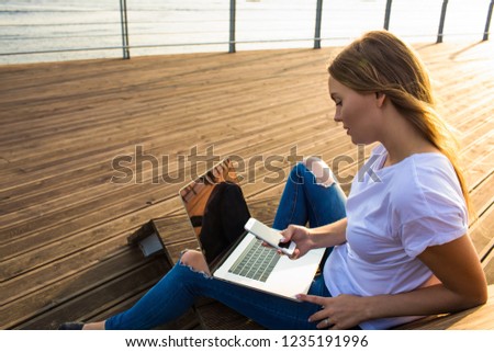 Fashionable hipster girl checking notifications on smartphone during online learning via laptop computer while sitting on embankment in sunny summer evening. Female sending sms via cellphone 