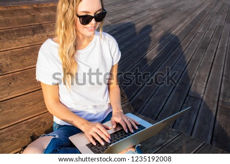 Hipster girl in fashionable sunglasses keyboarding on laptop computer preparing  article for web public while sitting outdoors on wooden bench near copy space for promotional content.