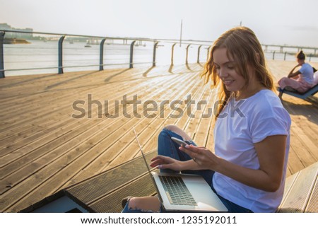 Smiling woman online shopping store via mobile phone after distance work on laptop computer, sitting outdoors on promenade during beautiful summer evening. Female reading lifestyle blog on cellphone 