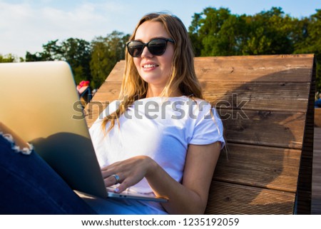 Young woman clever publication specialist typing new advertising article on laptop computer while sitting in park in sunny summer day. Stylish hipster girl having online video call via notebook 