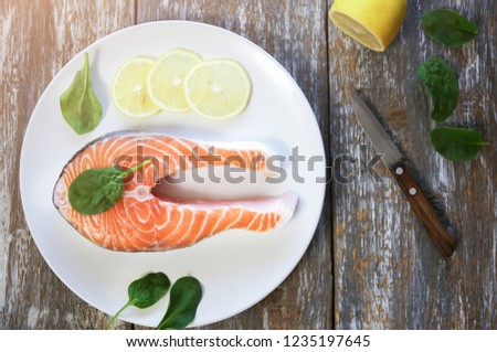 A piece of fresh salmon fish steak on a white plate with spinach and lemon, on  a gray wooden background. Omega 3 vitamin, healthy lifestyle. Natural vegetarian food photo. Top view.
