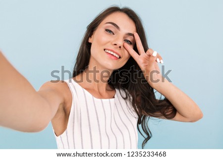 Image of beautiful excited young woman isolated over blue background wall make a selfie by camera showing peace gesture.