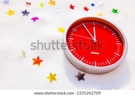 Red wall clock isolated on white background. The clock shows the last minutes before the new year. The clock lies on the snow with stars.