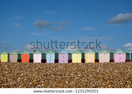 A row of ten colorful beach huts are in a line in the centre of the image in the foreground is a pebble beach above is a big blue sky with white clouds the sun is shining, Brighton, Sussex, UK. Royalty-Free Stock Photo #1235290201