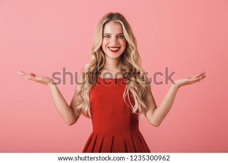 Portrait of a smiling young woman in red dress standing isolated over pink background, presenting copy space