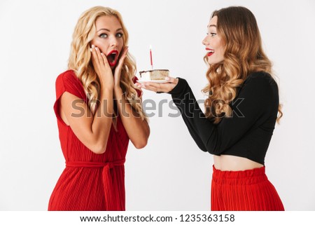 Portrait of two happy young smartly dressed women wearing makeup standing isolated over white background, celebrating together with piece of a cake