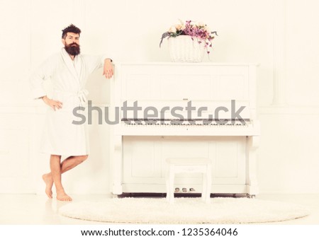 Man with beard in bathrobe enjoys morning while standing near piano. Talented musician concept. Man serious stands and leans on piano musical instrument in white interior on background.