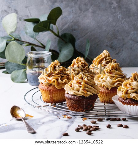 Fresh baked homemade cupcakes with coffee buttercream and caramel standing on cooling rack with eucalyptus branch and coffee beans above over white marble kitchen table. Square image