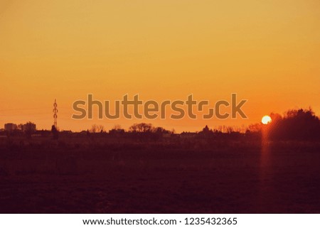 Sunset landscape, orange sky, sun setting down, dark horizon with silhouettes of electricity pylon, wires, houses and trees, rural countryside