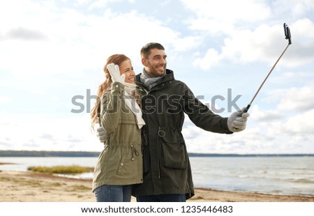 technology and people concept - smiling couple taking picture by smartphone on selfie stick on beach in autumn