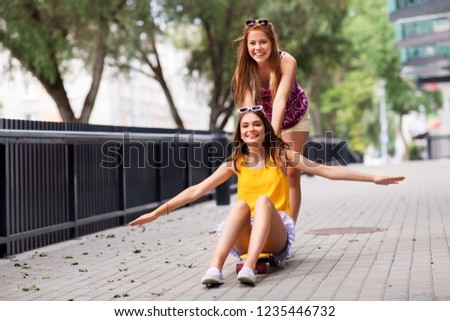 friendship, leisure and people concept - happy teenage girls or friends riding skateboard on city street in summer