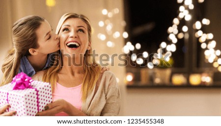 people, holidays and family concept - happy daughter giving birthday present and kissing her mother at home over garland lights background