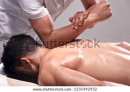 side view, two young man, 20-29 years old, sports physiotherapy indoors in studio, photo shoot. Strong Physiotherapist hard massaging relaxed patient neck back side, with his elbow.