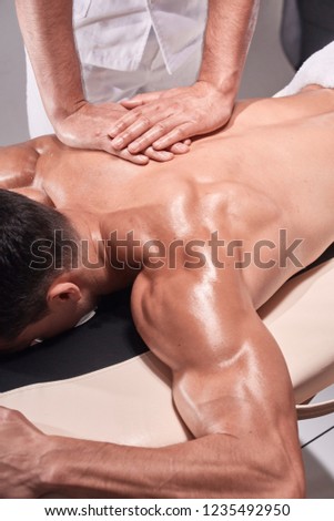 two young man, 20-29 years old, sports physiotherapy indoors in studio, photo shoot. Physiotherapist massaging muscular patient back with his hands close-up.