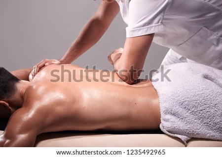 side view, two young man, 20-29 years old, sports physiotherapy indoors in studio, photo shoot. Physiotherapist massaging muscular patient lower back with his hands.