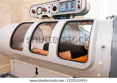 Treatment in an oxygen capsule. A beautiful girl in a black T-shirt and white pants is lying in a pressure chamber. The concept of modern medicine Royalty-Free Stock Photo #1235552455