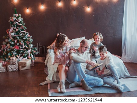Happy family sitting on the floor by the bed in the new year interior. Dad is reading a book. Children laugh. Cozy room. Warm tone.