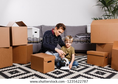 Picture of happy father and son with microscope sitting on floor among cardboard boxes