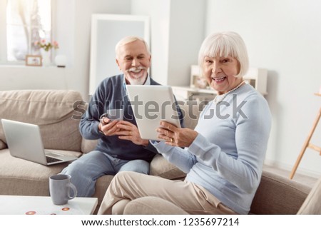 Happy smile. Beaming elderly woman wearing blouse smiling bright while showing pictures on the gadget