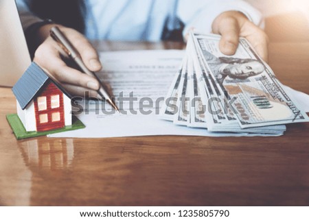 Estate agent with house model holding money and pointing the pen on document showing where to sign.